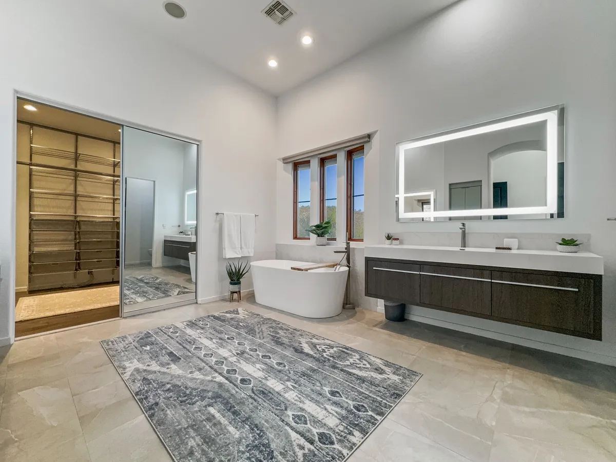 A spa-like bathroom with a freestanding bathtub, a large backlit mirror, and modern cabinetry. Natural light streams in from a nearby window, adding to the tranquil, luxurious atmosphere ideal for relaxation and rejuvenation.
