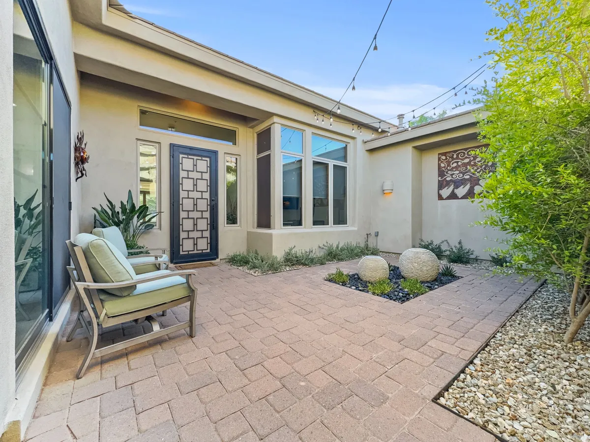 A peaceful exterior seating area near a decorative entrance door, surrounded by landscaped stones and shaded by trees. String lights create a magical ambiance, ideal for evening socializing or enjoying quiet mornings outdoors.