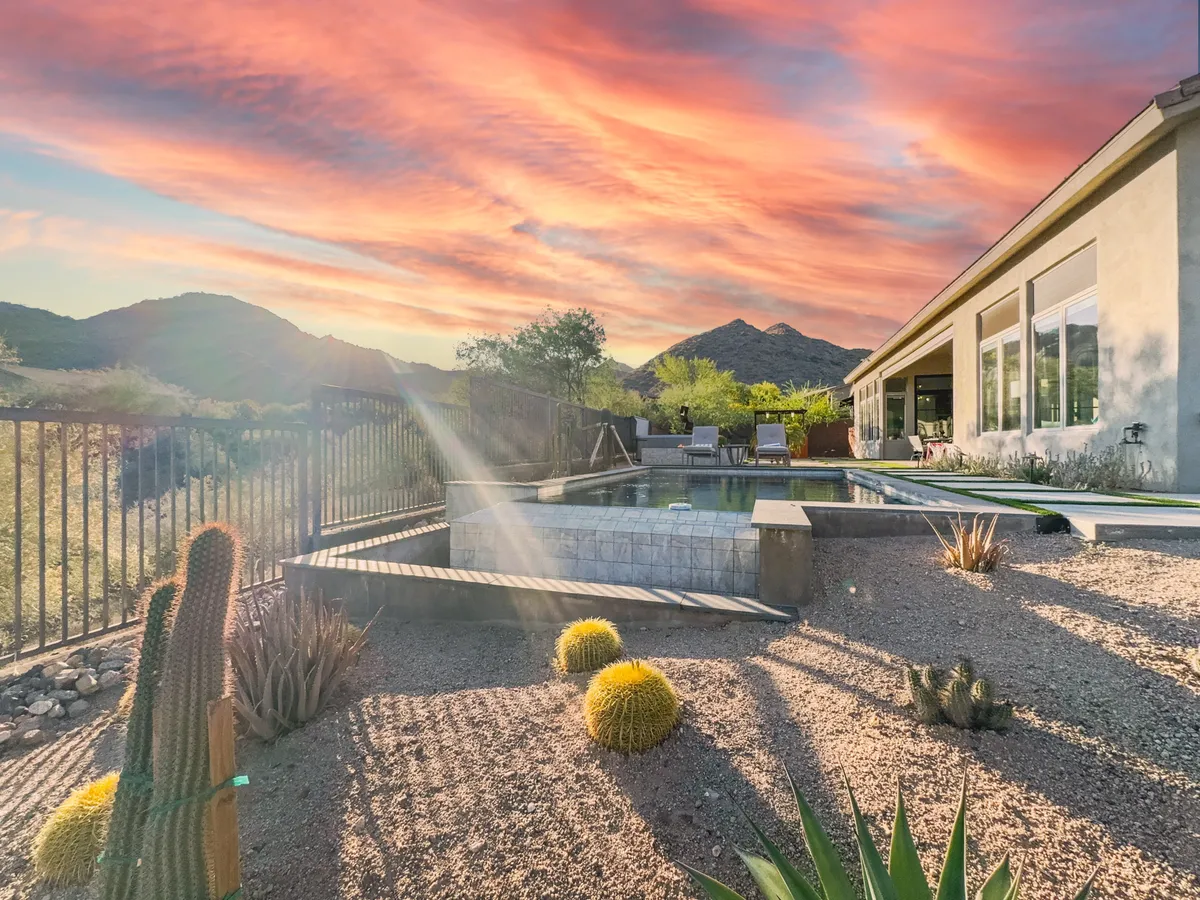 This exterior image features a sparkling swimming pool surrounded by desert landscaping, with striking cactus plants in the foreground and the McDowell Mountains rising in the background beneath a dramatic sunset sky. The setting exudes a sense of direct connection to nature and the surrounding trails.