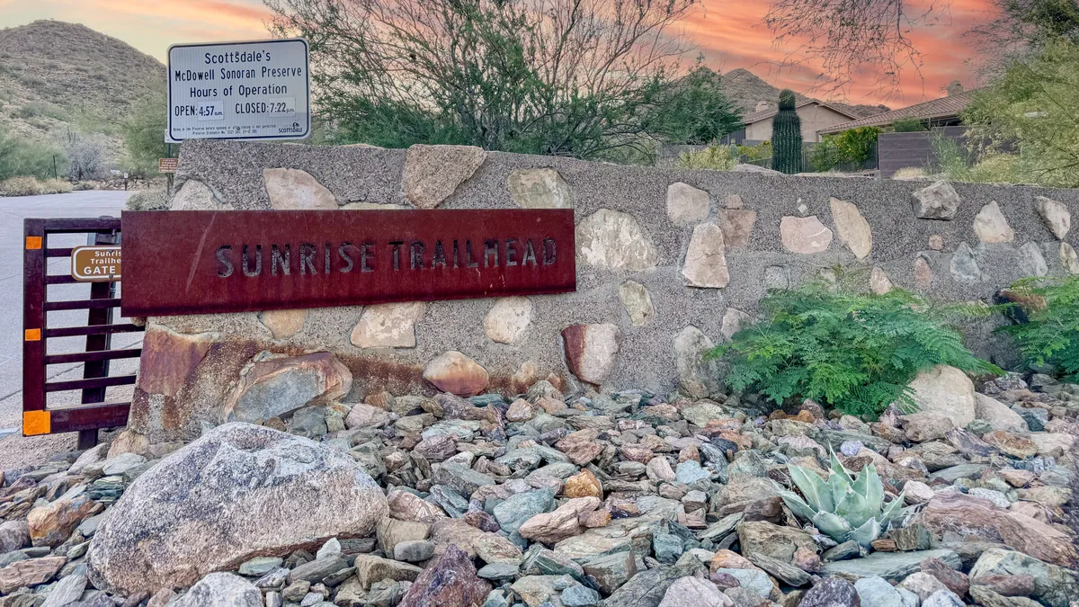 The Sunrise Trailhead sign stands against a rocky desert landscape with local vegetation, capturing the spirit of adventure and direct access to natural trails.