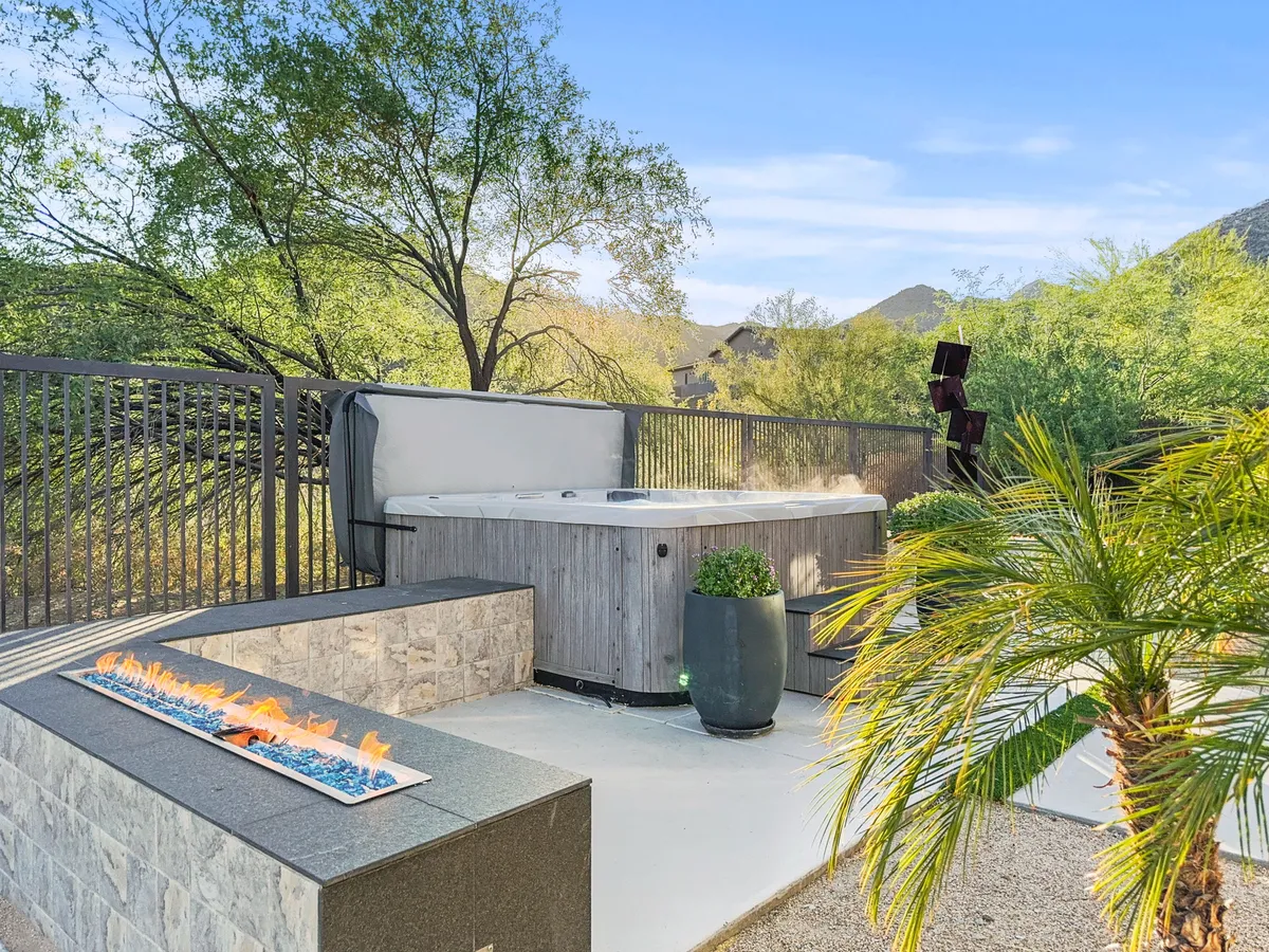 An exterior space featuring a bubbling hot tub, a dramatic fire feature, and landscaped surroundings with palm plants. The backdrop includes mountains and an open sky, evoking a serene, luxurious atmosphere.