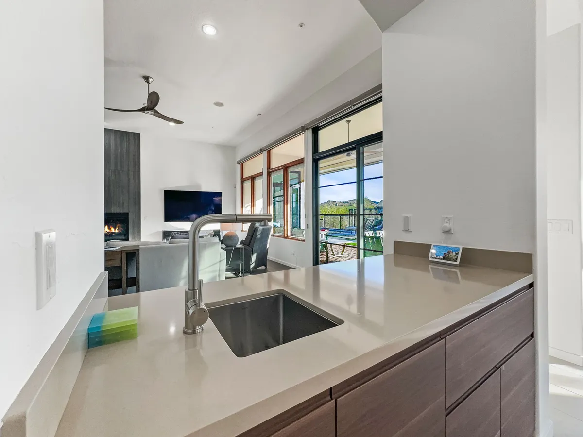 A contemporary, open-plan kitchen and living space. The kitchen sink and countertop overlook a spacious living area, with large windows providing sunrise views and abundant natural light. A fireplace adds warmth while the open flow encourages both relaxation and entertaining.