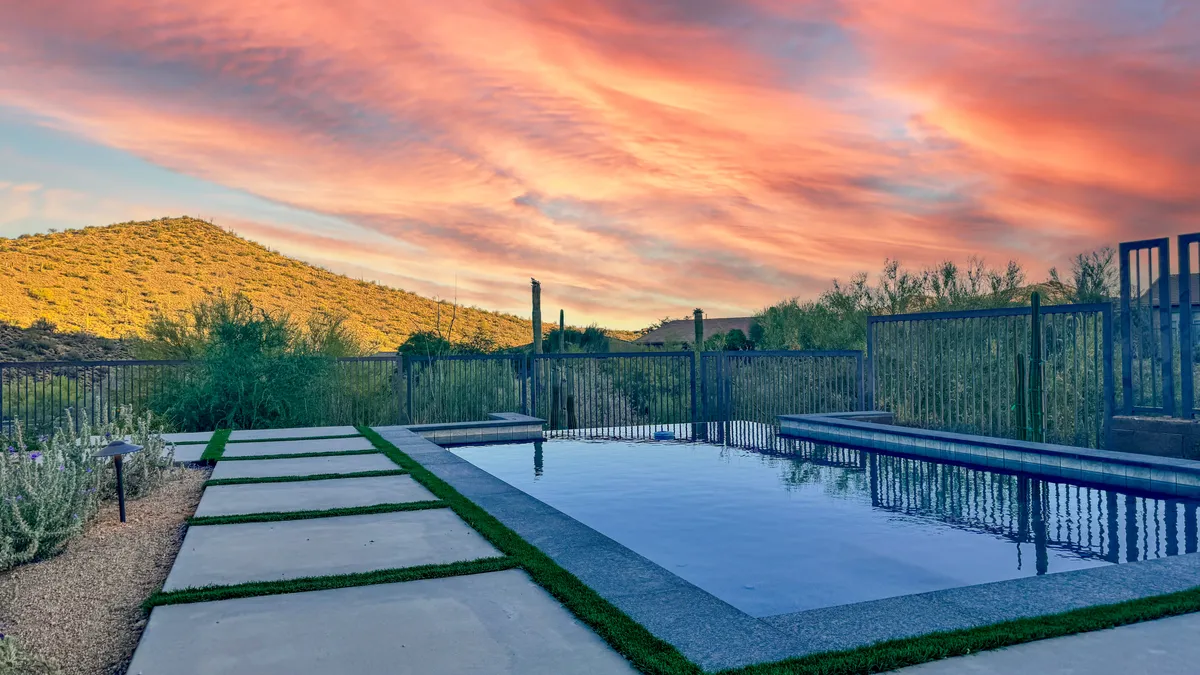 A tranquil backyard scene featuring a sparkling swimming pool set against a backdrop of mountains and a colorful sunset sky. The pool area is framed by subtle landscaping and offers plenty of space for both relaxation and play.