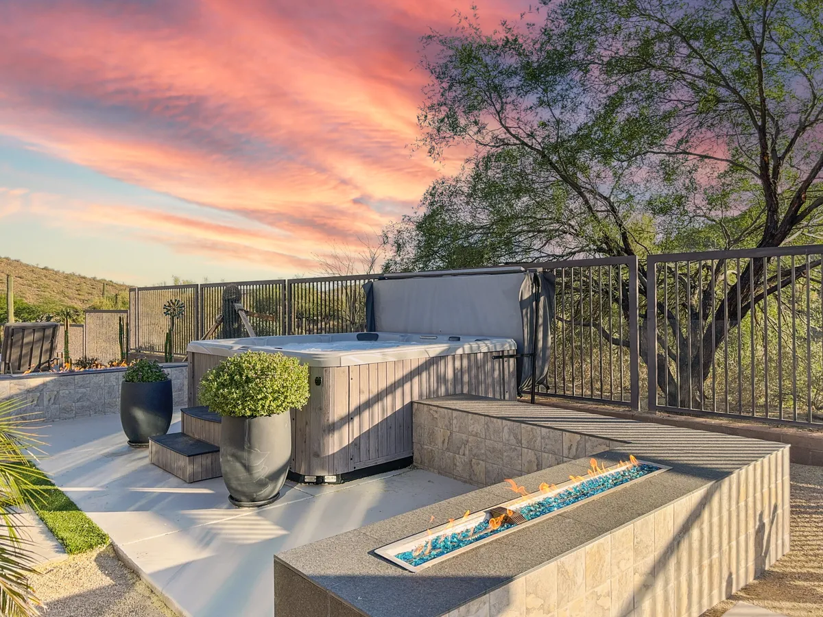 This exterior image features a luxurious hot tub tucked beside a modern fire feature, surrounded by planters. The setting is tranquil and perfect for unwinding, with the warm glow of sunset adding to the atmosphere. The design cues suggest a private, resort-style experience in the comfort of home.