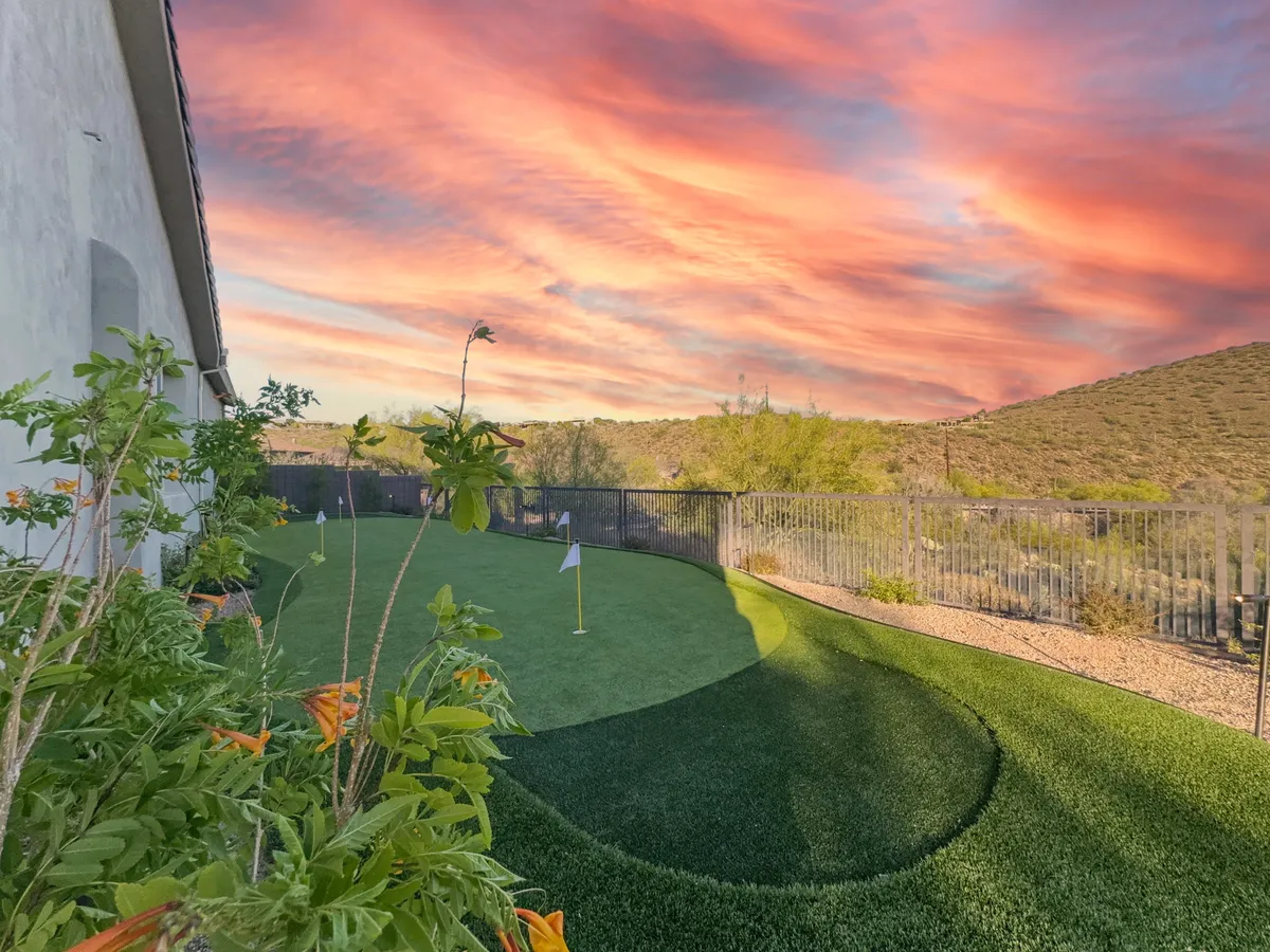 An exterior shot of a vibrant putting green nestled in lush greenery, with a fenced yard and a dramatic sunset sky overhead. The scene feels playful and serene, suggesting activities like family golf competitions or peaceful sunset moments.