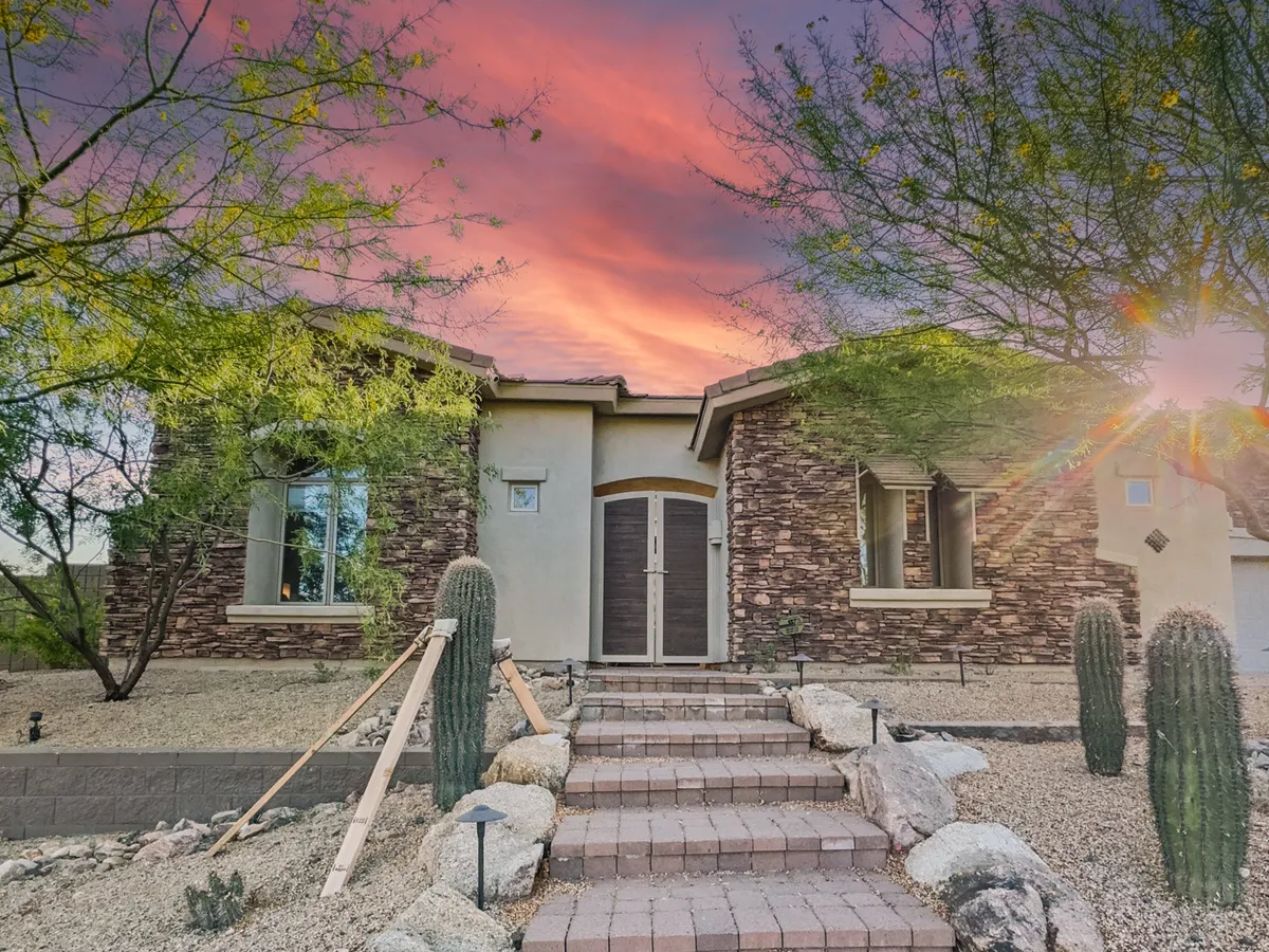 A welcoming entryway framed by natural stone, cacti, and desert plants, with stairs leading up to double doors. The entrance is set against the backdrop of Arizona’s iconic landscape, suggesting both hospitality and easy access to the great outdoors.