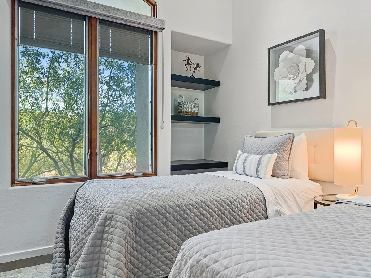A bedroom showcasing two beds with gray quilts, a large sunlit window, decorative shelving, and framed artwork. The shelving unit and neutral color palette create a cozy, adaptable environment ideal for guests seeking either twin or king setups.