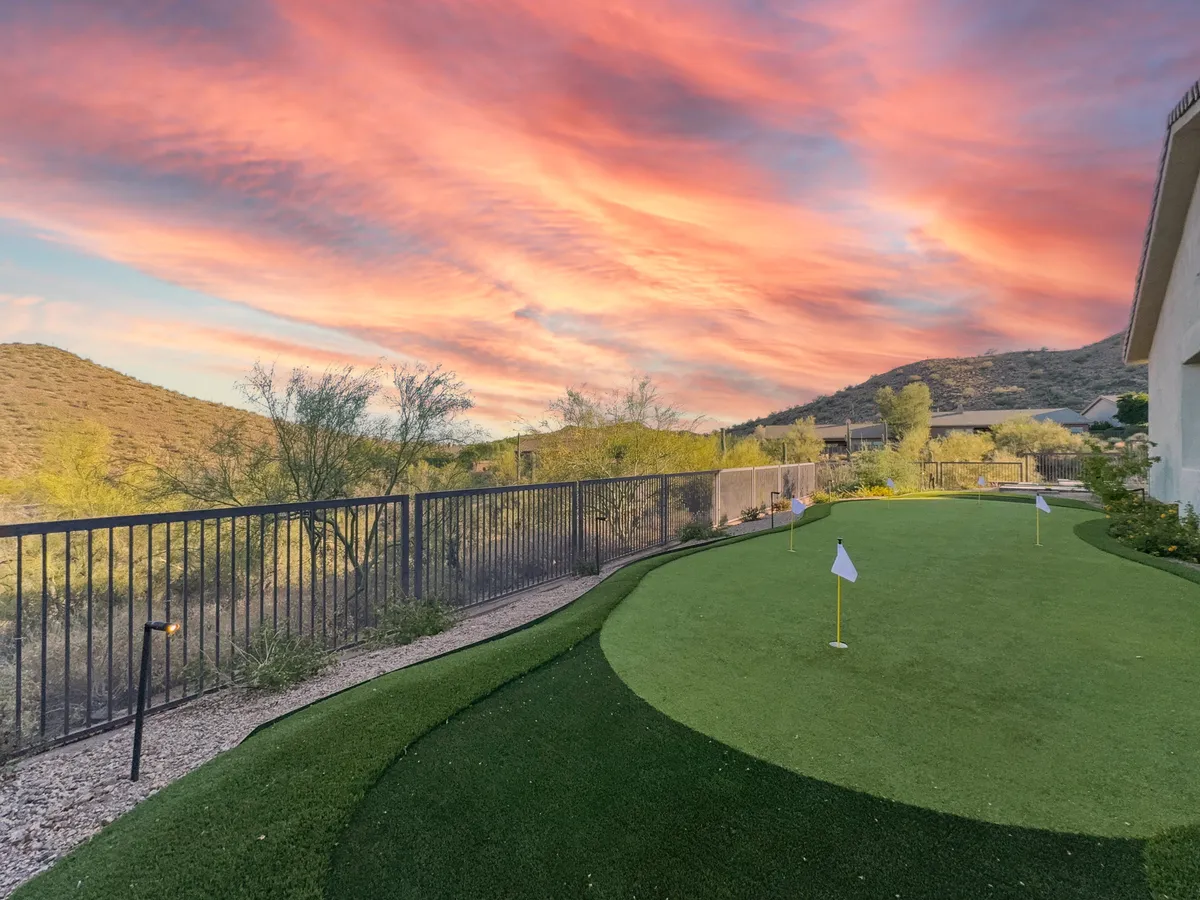A lush golf putting green set against a mountainous backdrop, under a colorful sunset sky. The space is bordered by greenery and fencing, offering both recreation and tranquility.